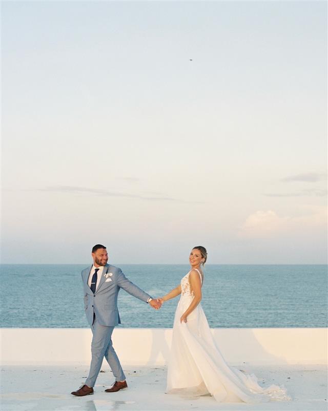 Teya and Charles holding hands on a white terrace by the sea at their destination wedding under a clear sky.