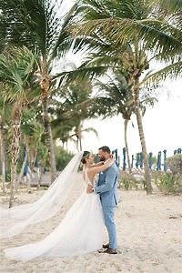 Teya and Charles embrace on a sandy beach with palm trees at their romantic destination wedding.