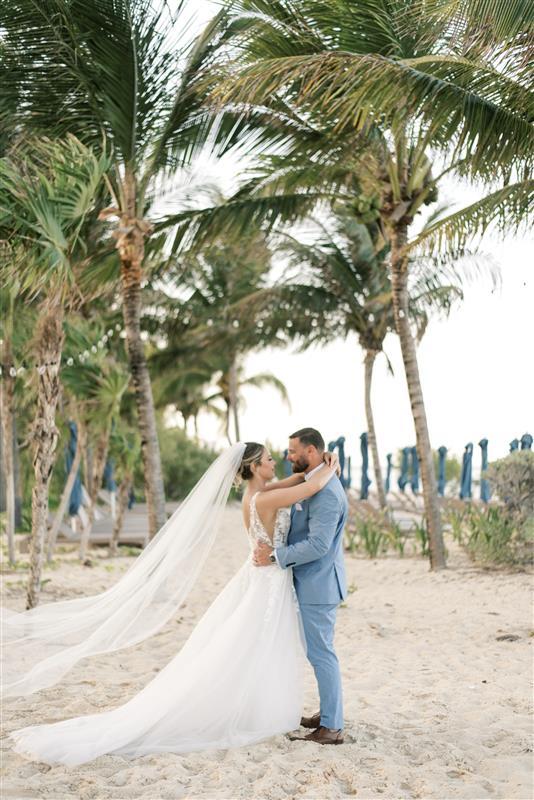 Teya and Charles embrace on a sandy beach with palm trees at their romantic destination wedding.