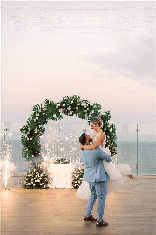 Groom lifts bride on deck beneath floral arch at destination wedding, sparklers light by the ocean.