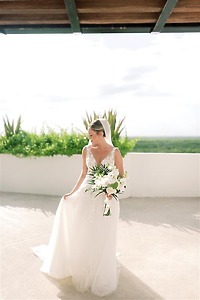 Bride in white dress holding a bouquet stands at a destination wedding under a shaded outdoor structure.