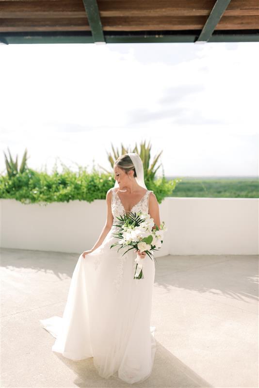 Bride in white dress holding a bouquet stands at a destination wedding under a shaded outdoor structure.