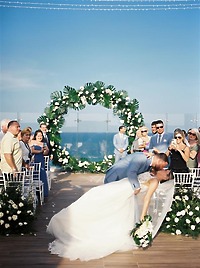 Teya and Charles share a kiss beneath a floral arch at their destination wedding as guests watch.