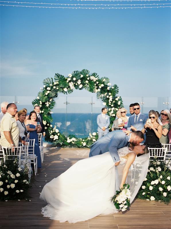Teya and Charles share a kiss beneath a floral arch at their destination wedding as guests watch.