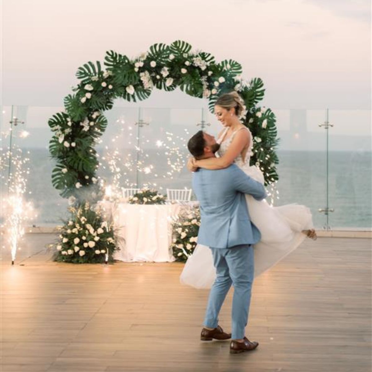 Groom lifts bride by floral arch with sparklers at destination wedding on waterfront.