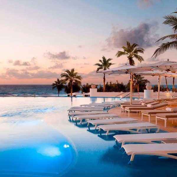 Infinity pool with wedding décor, lounge chairs, and umbrellas overlooks the ocean at sunset, palms nearby.