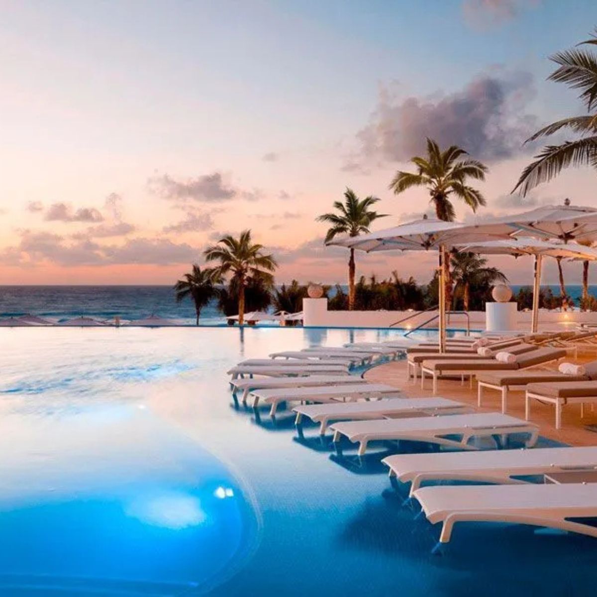 Infinity pool with wedding décor, lounge chairs, and umbrellas overlooks the ocean at sunset, palms nearby.