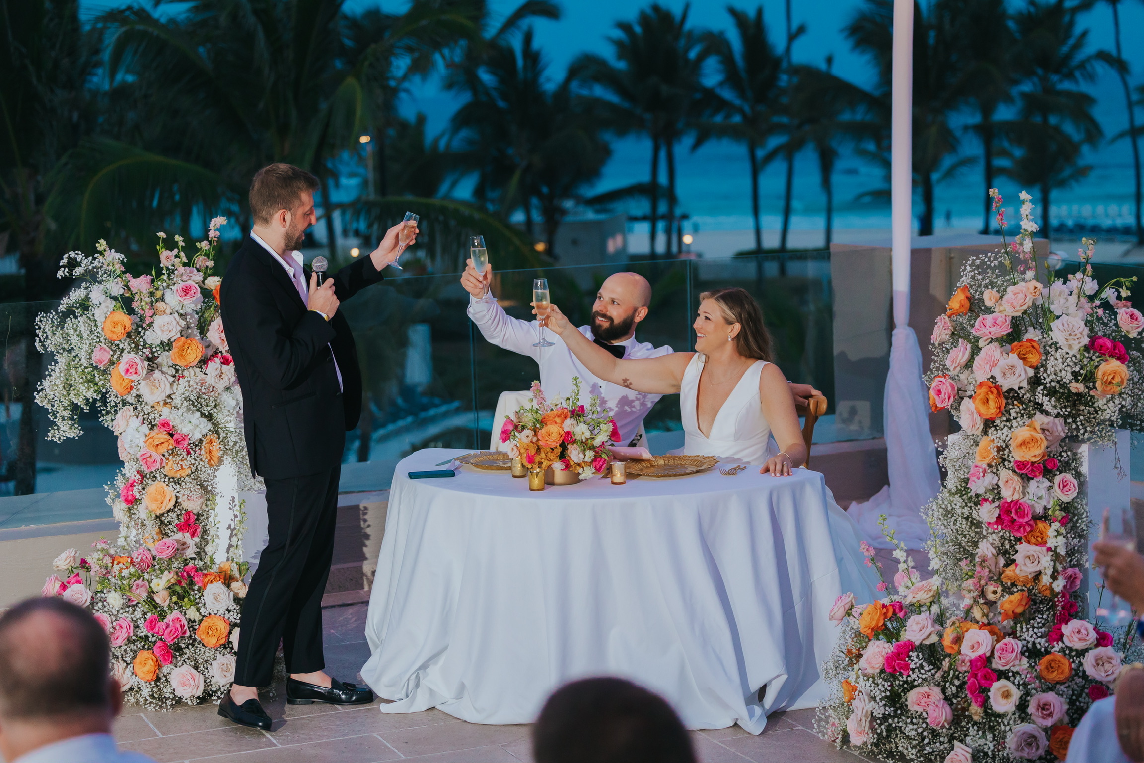 A man raises a toast to Billi and Trevor at their decorated outdoor destination wedding at dusk.