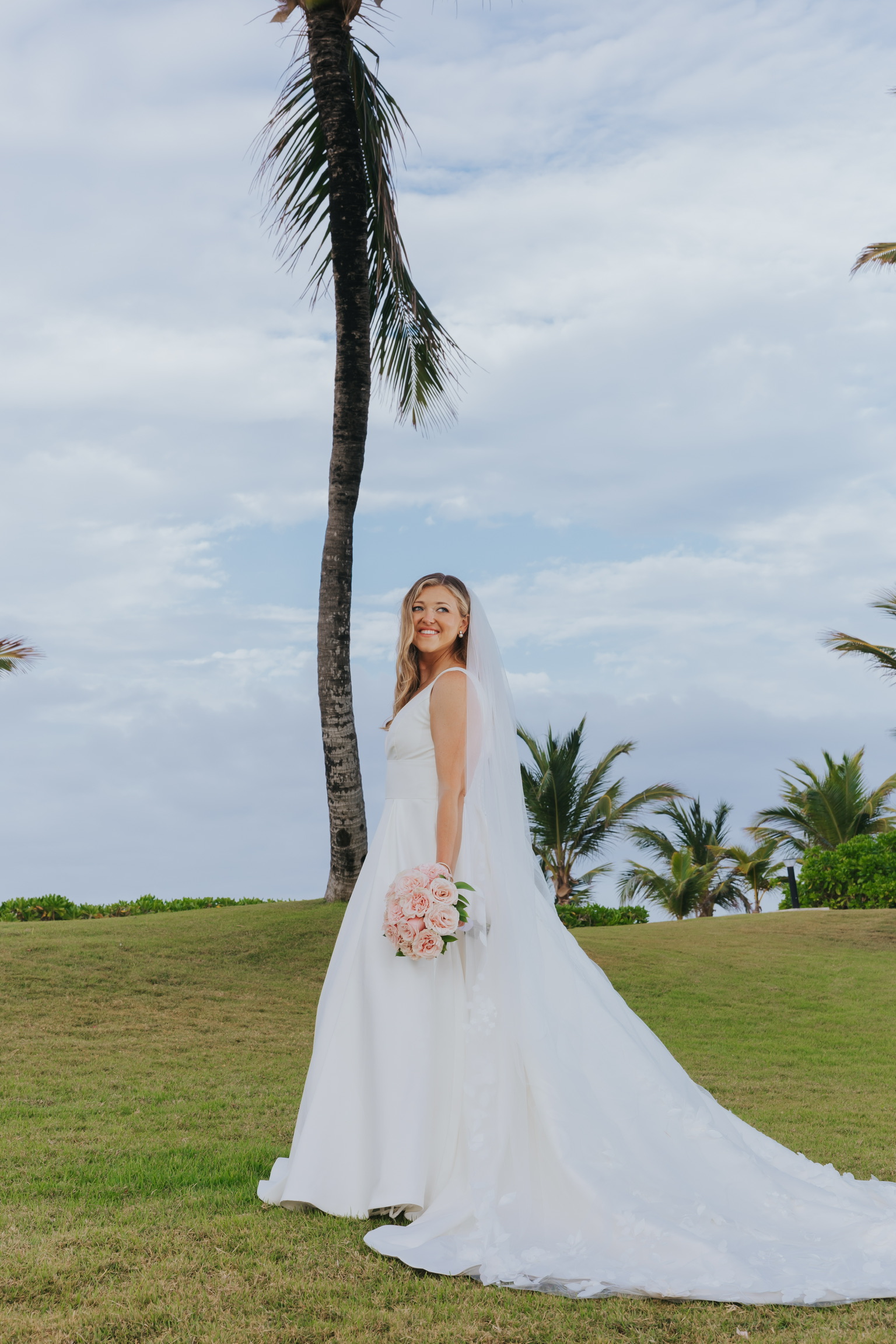 Bride in white wedding gown holding bouquet stands on grassy lawn near palms—Billi and Trevor's destination wedding.