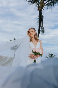Bride in "Billi and Trevor (Copy)" dress holds pink bouquet at a destination wedding with palm trees.