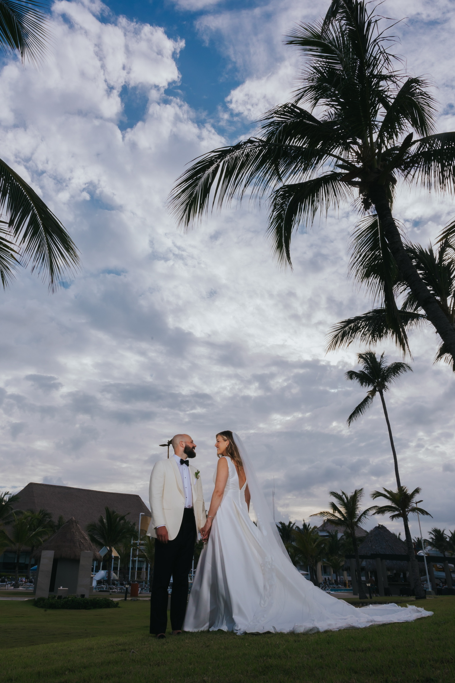 Billi and Trevor (Copy) hold hands outdoors under palm trees at a destination wedding beneath cloudy skies.