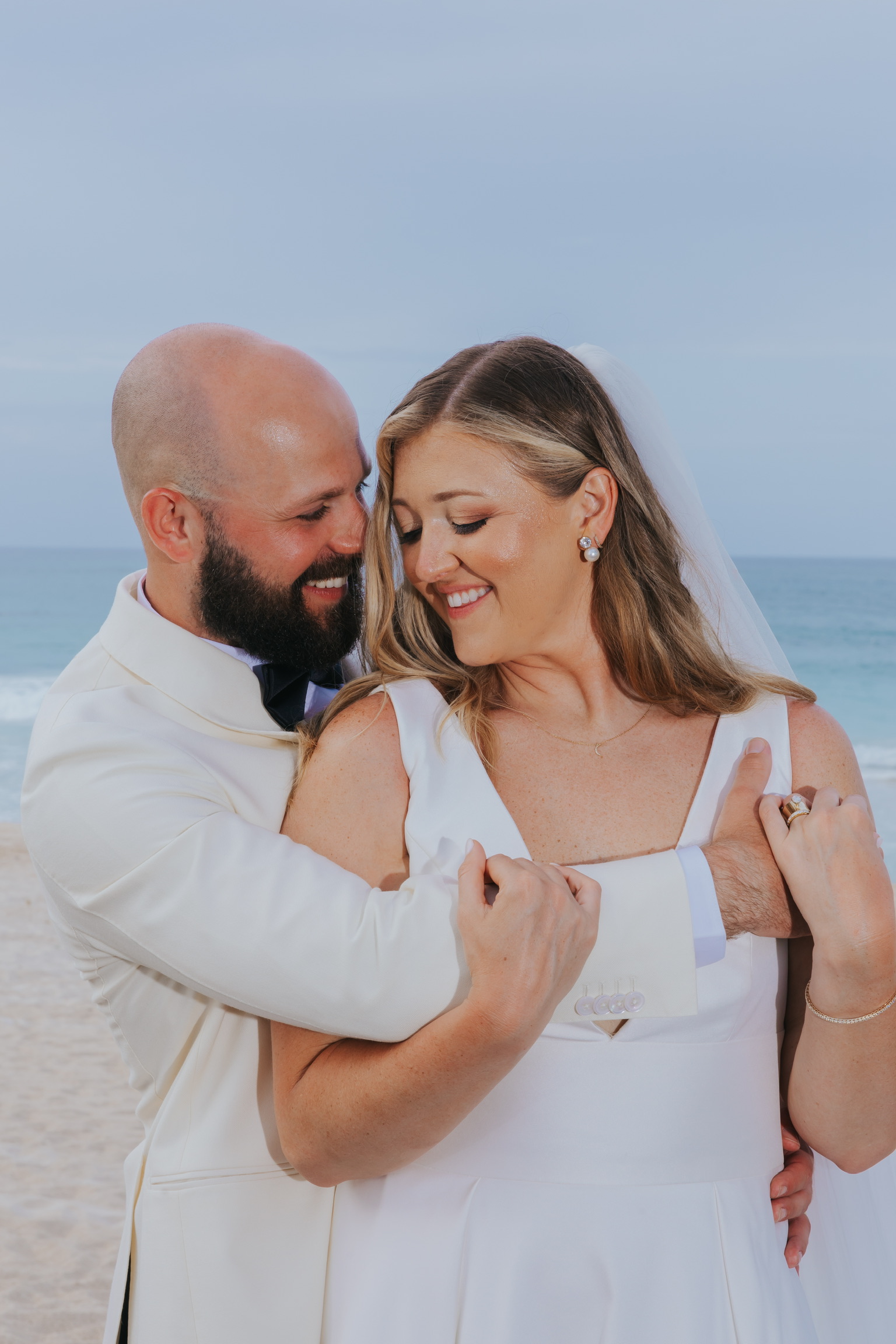 Billi and Trevor embrace and smile on a beach during their destination wedding, ocean waves behind them.