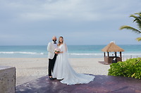 Billi and Trevor pose on a beach at their destination wedding, with the ocean and a hut behind them.