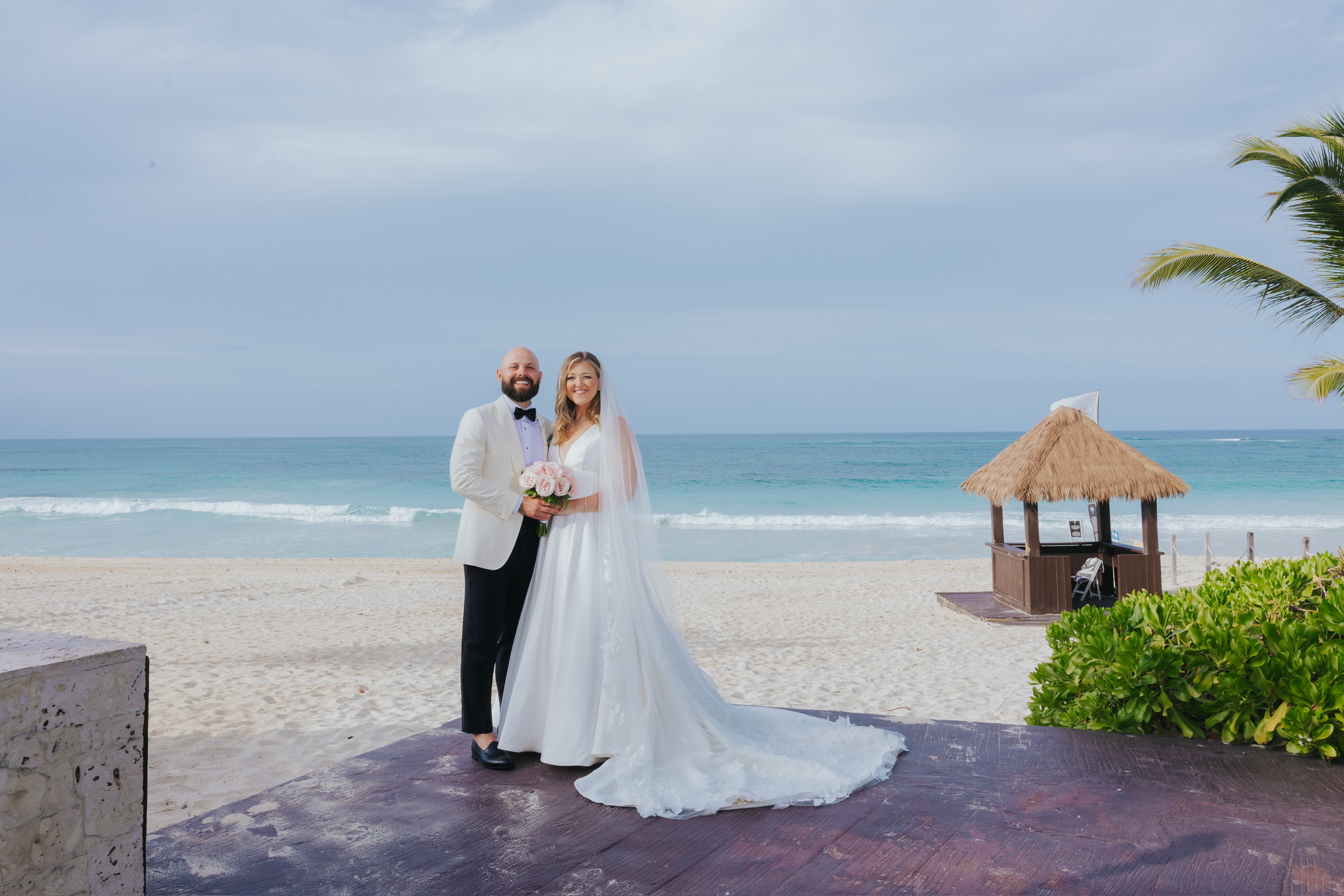 Billi and Trevor pose on a beach at their destination wedding, with the ocean and a hut behind them.
