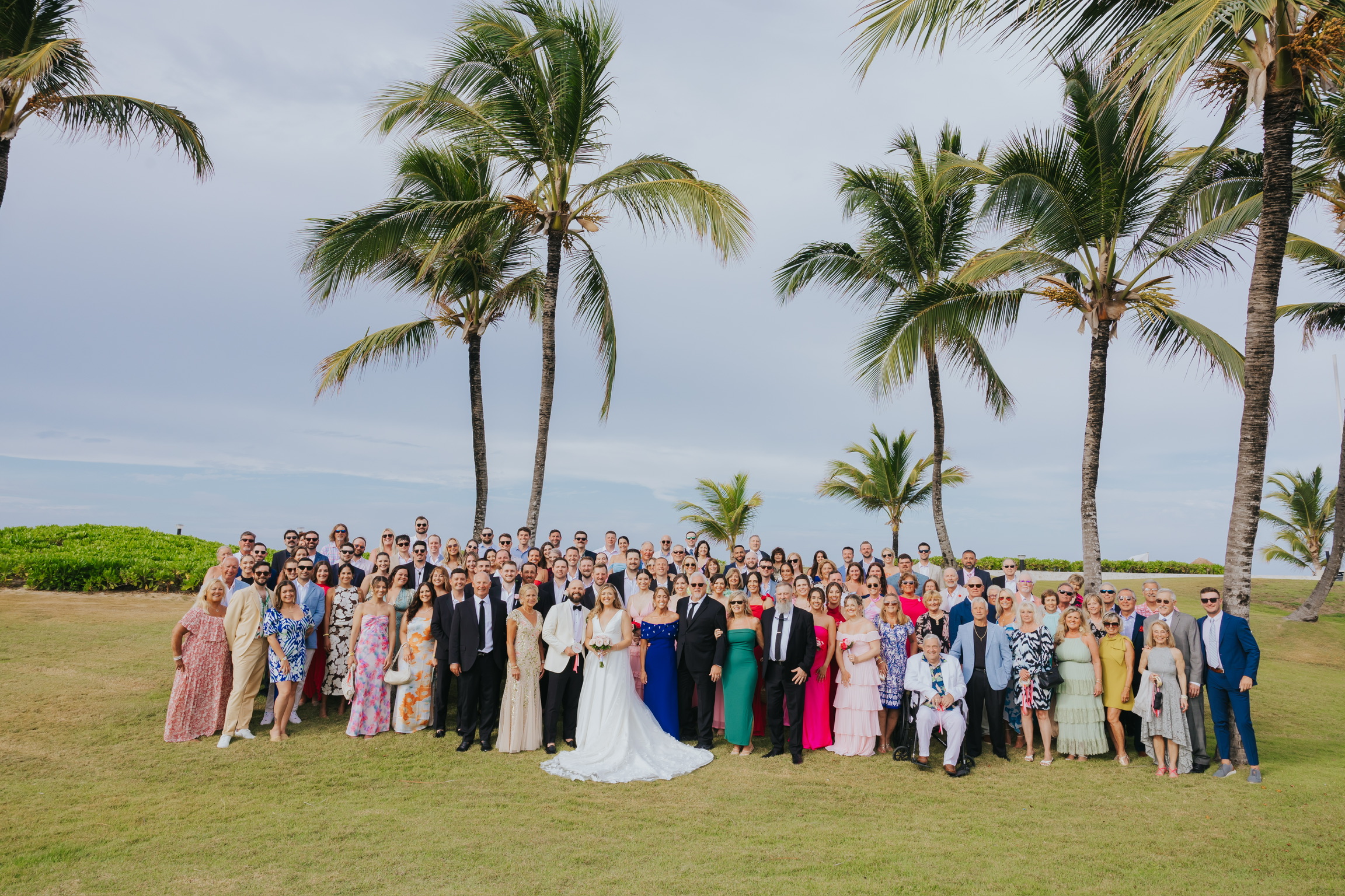 A destination wedding group outdoors under palm trees, with Billi and Trevor in the center.