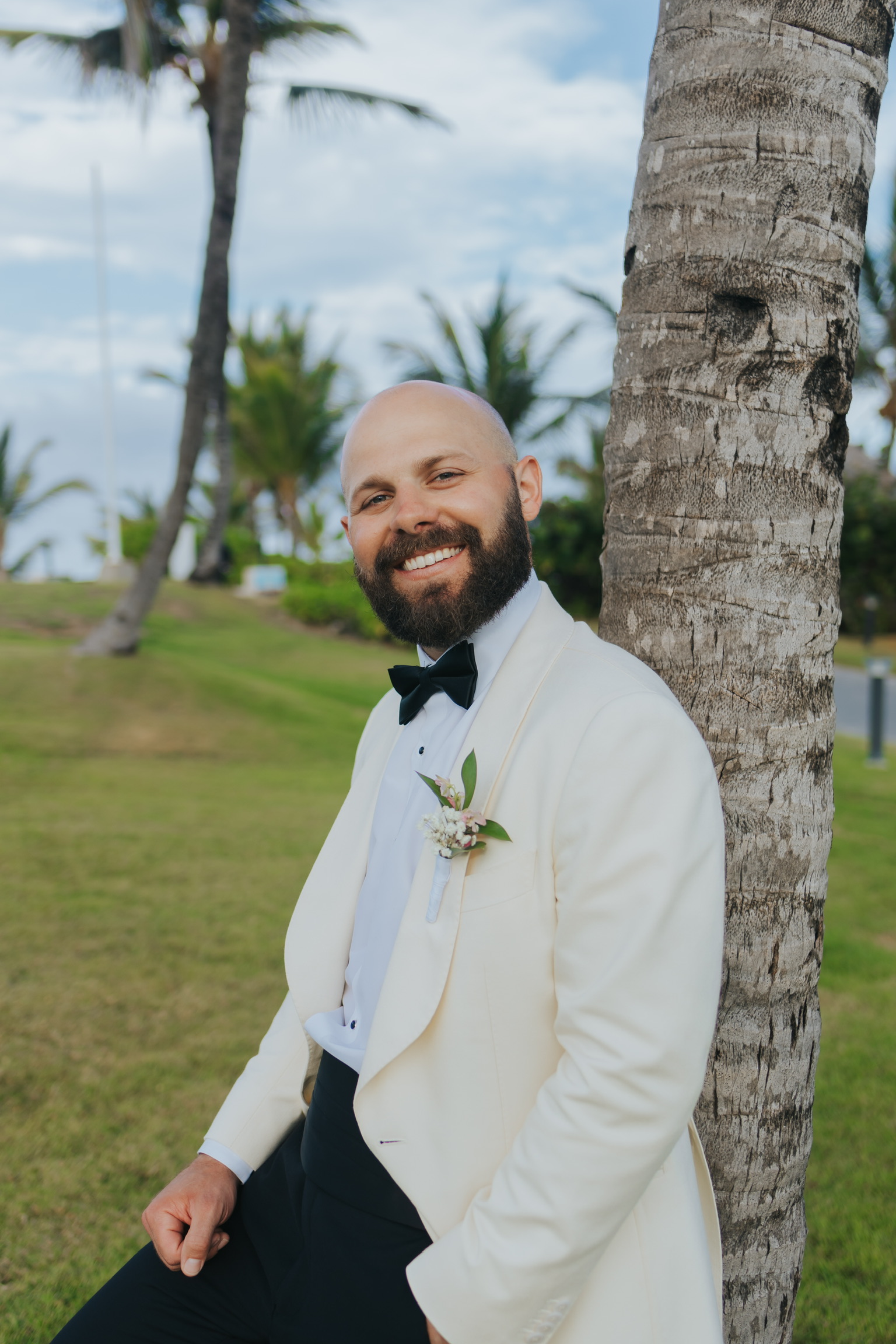 Man in a Billi and Trevor tuxedo leans on a palm tree, smiling at a destination wedding.