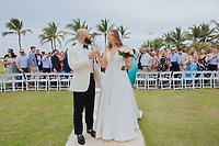 Billi and Trevor walk down the outdoor wedding aisle as guests look on, framed by palm trees.