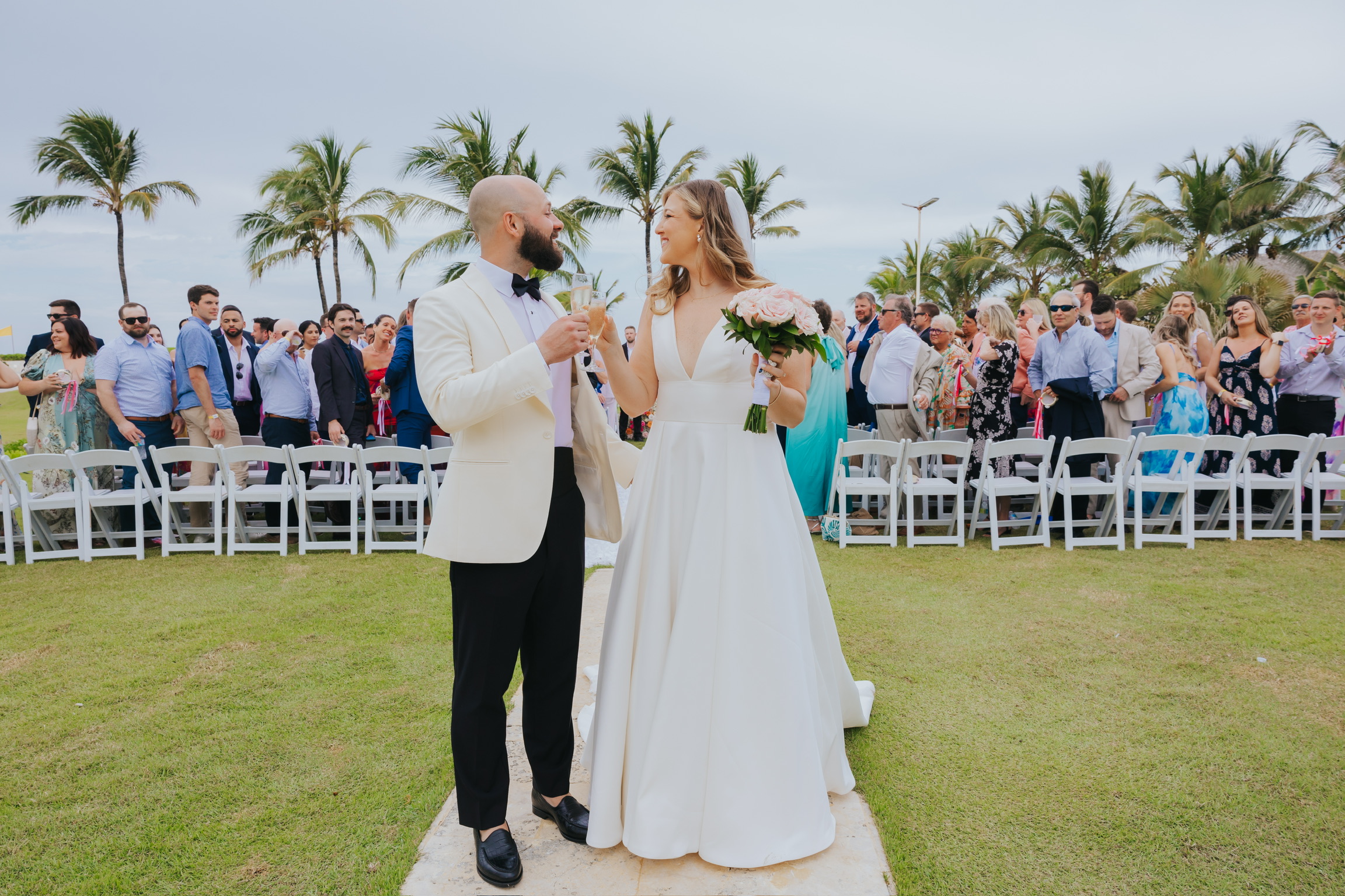 Billi and Trevor walk down the outdoor wedding aisle as guests look on, framed by palm trees.