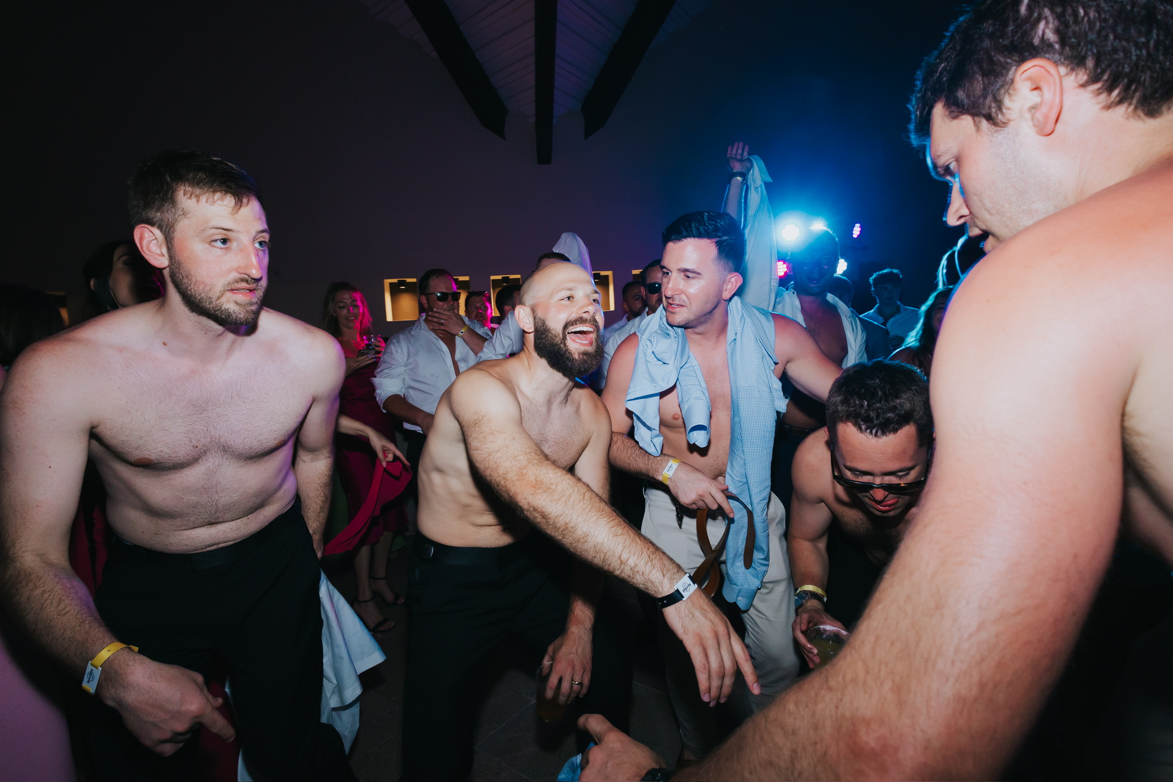 Shirtless men dance energetically at a lively indoor party during a Billi and Trevor destination wedding.
