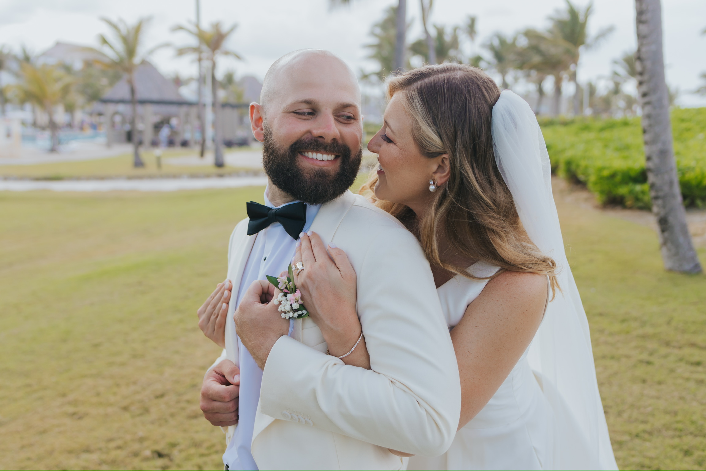 Billi and Trevor embrace and smile on the grass at their destination wedding with palm trees behind.
