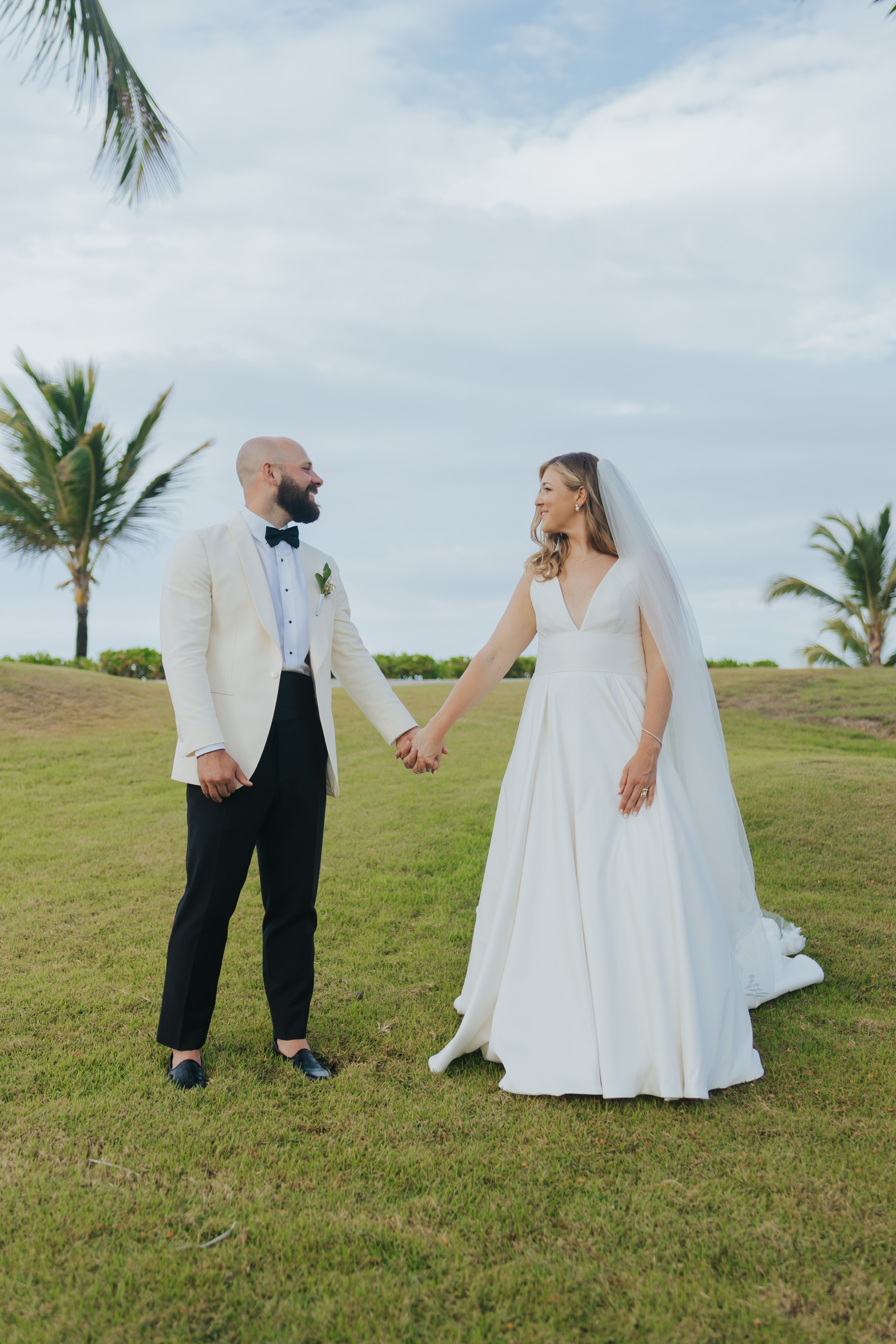 Billi and Trevor hold hands on the grass outdoors, gazing at each other during their destination wedding.