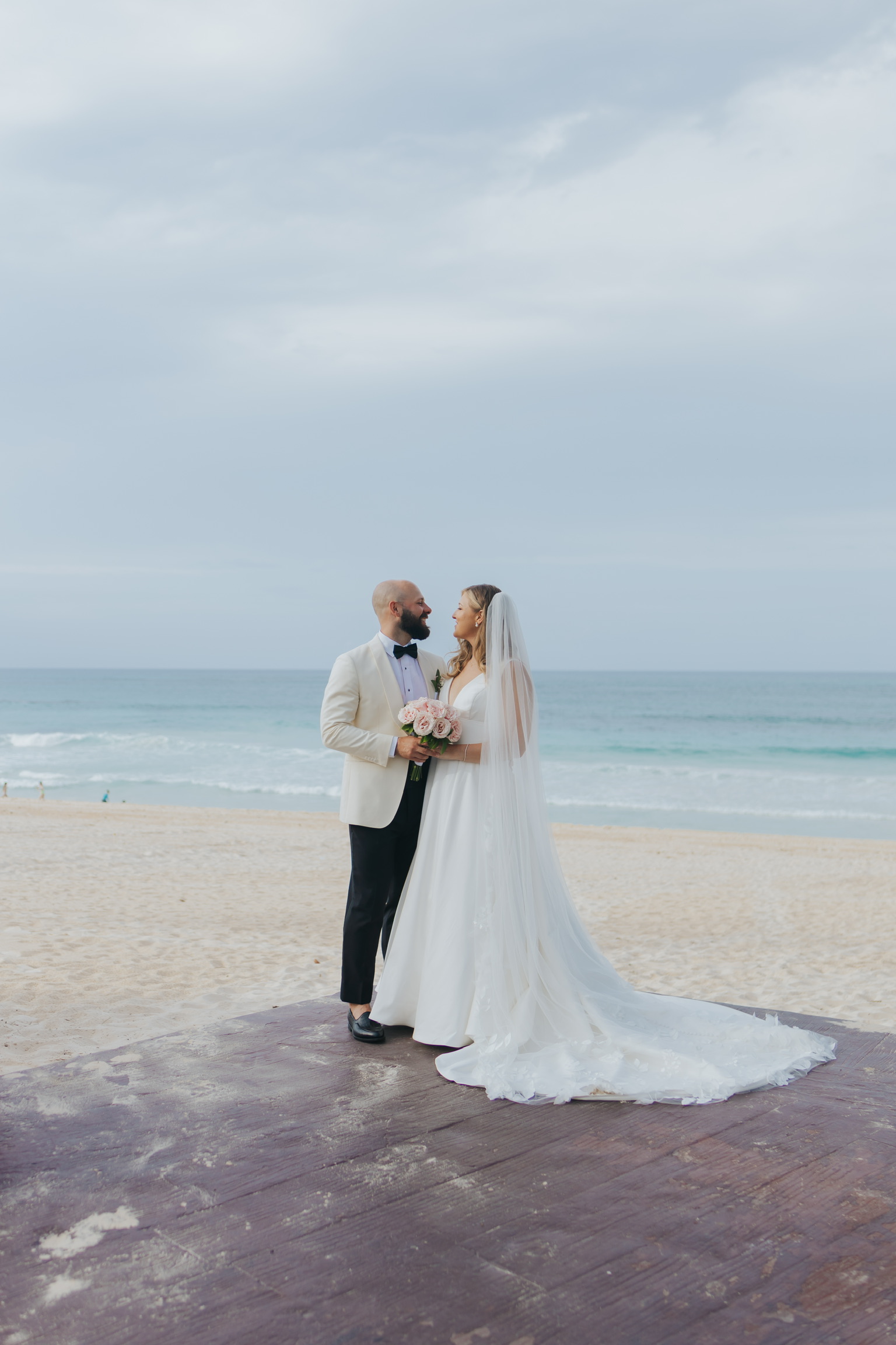 Billi and Trevor face each other on a beach, holding a bouquet at their destination wedding.