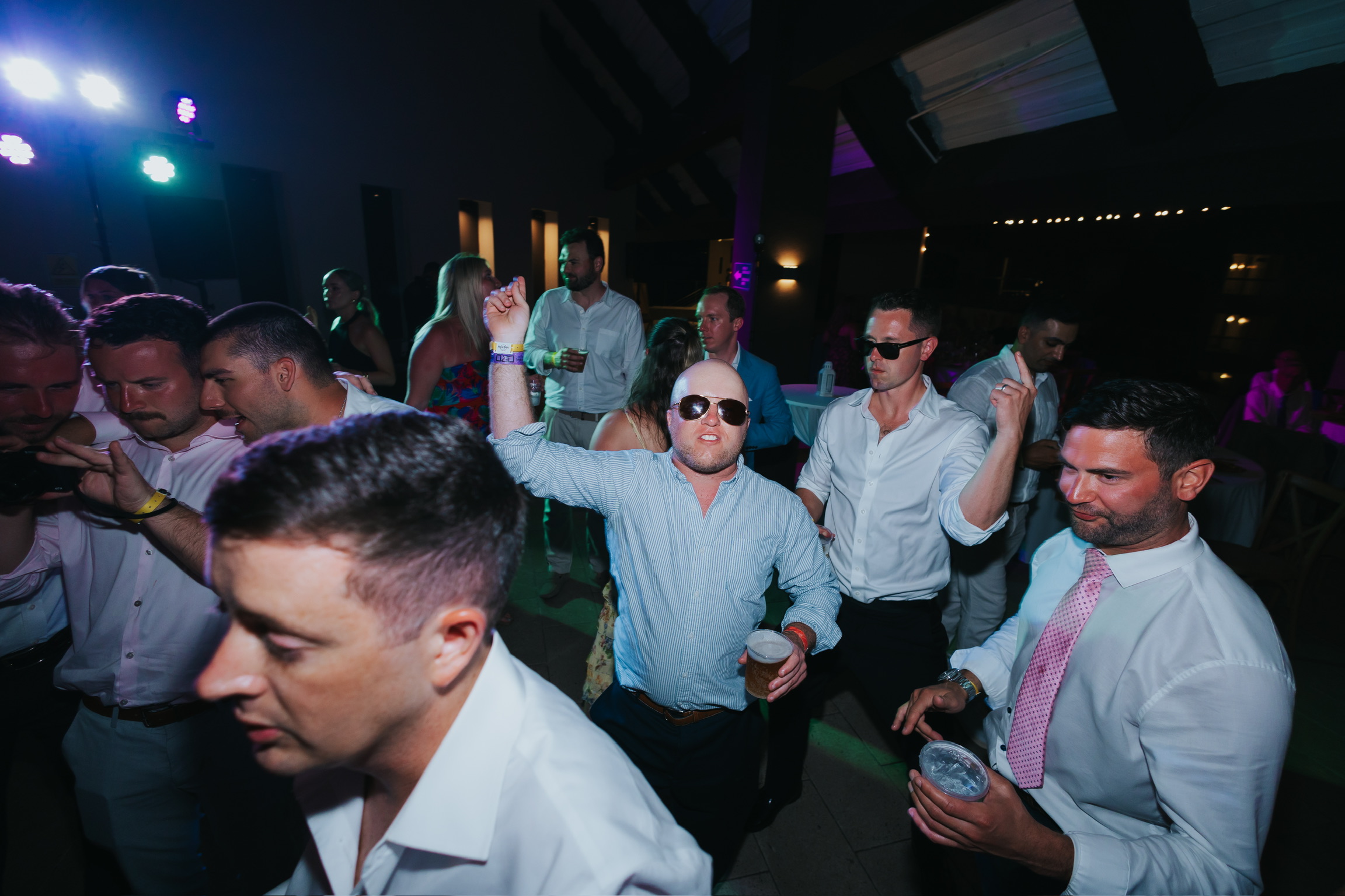 Men in dress shirts dance under vibrant lights at a Billi and Trevor destination wedding party.
