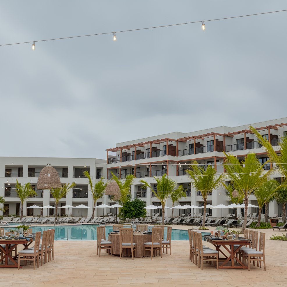 Outdoor dining tables set up by a pool for a destination wedding, with a modern hotel and palm trees nearby.