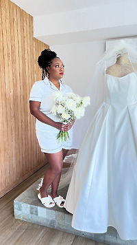 Woman in white pajamas holds a destination wedding bouquet by a wedding dress mannequin.