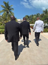 Three men in suits stroll a palm-lined path under blue skies at a stylish destination wedding.