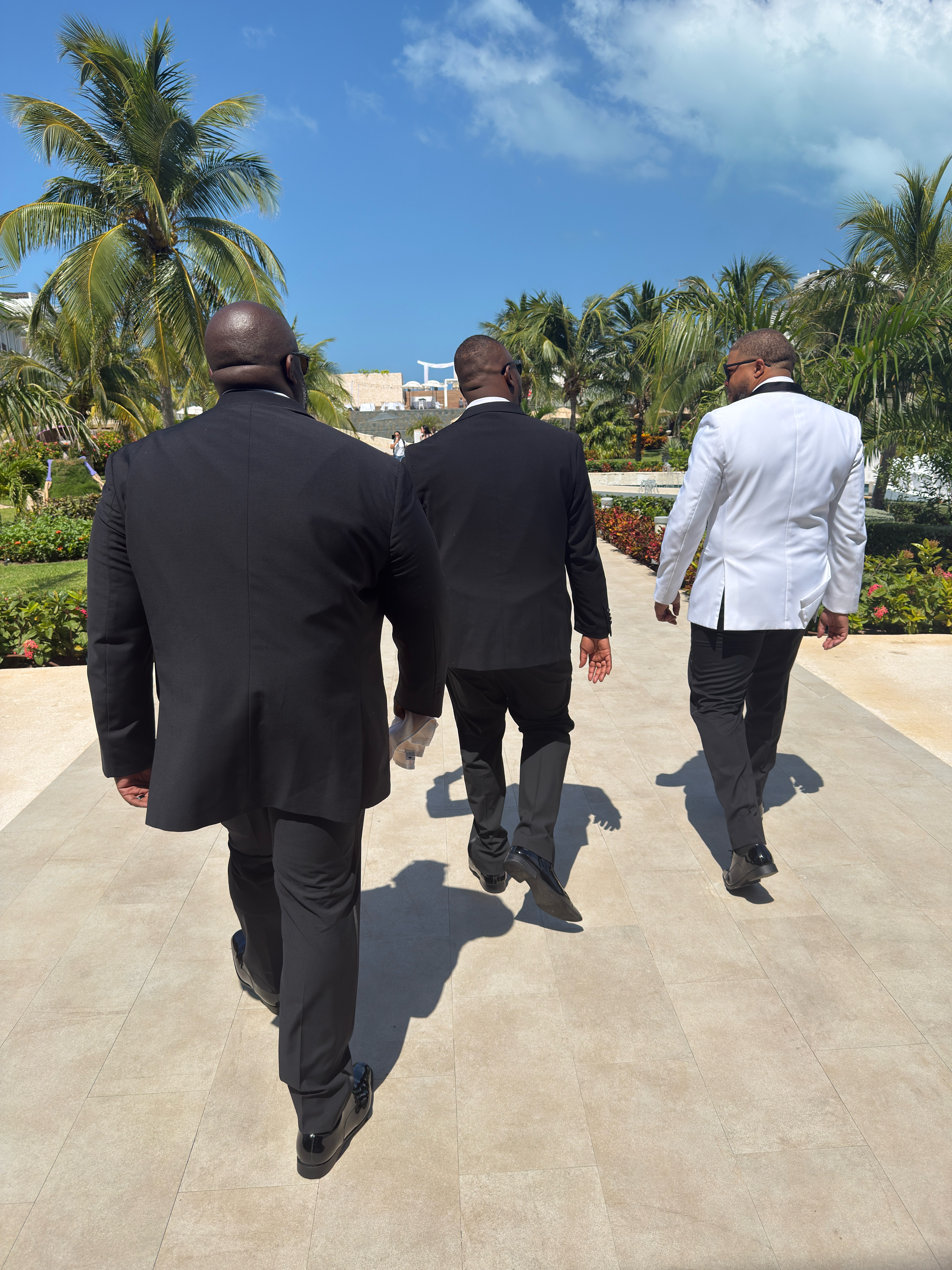 Three men in suits stroll a palm-lined path under blue skies at a stylish destination wedding.