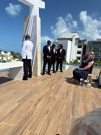 Groomsmen in suits stand at an outdoor wedding altar as guests sit under the sun at a destination wedding.