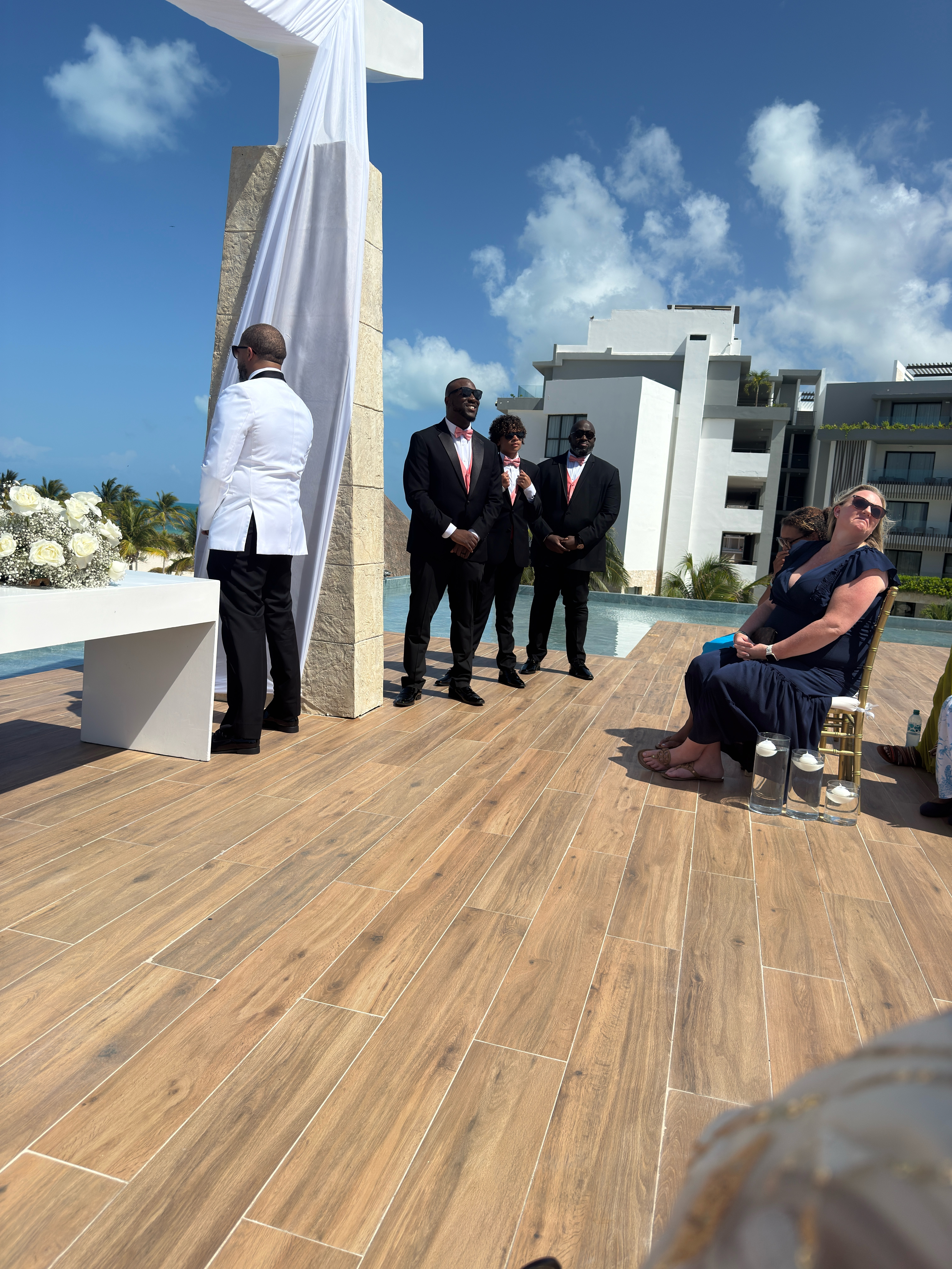 Groomsmen in suits stand at an outdoor wedding altar as guests sit under the sun at a destination wedding.