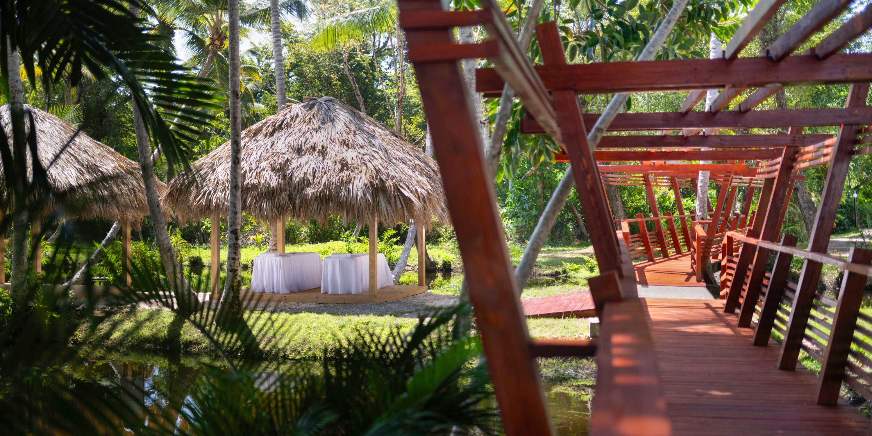 Wooden bridge leading to thatched huts with tables, ideal for a destination wedding in a tropical garden.