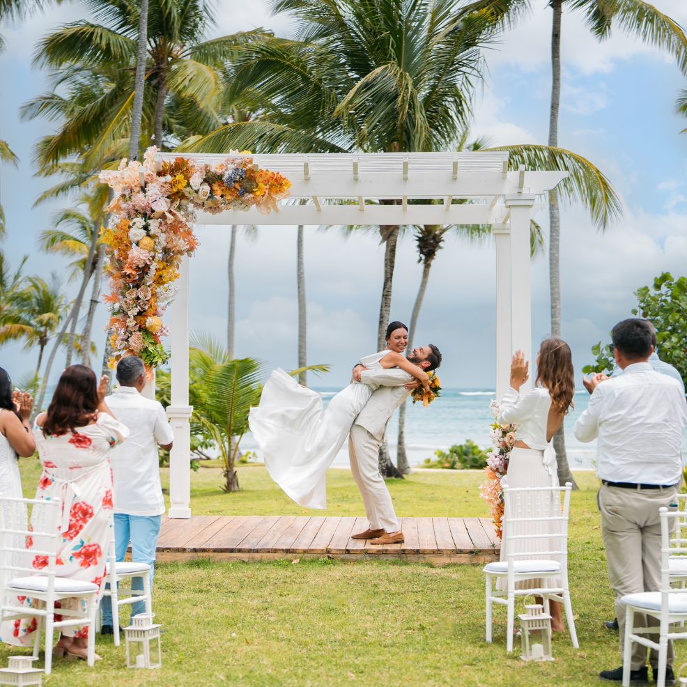 At a destination wedding, a groom lifts his bride beneath a floral arch as guests watch outdoors.