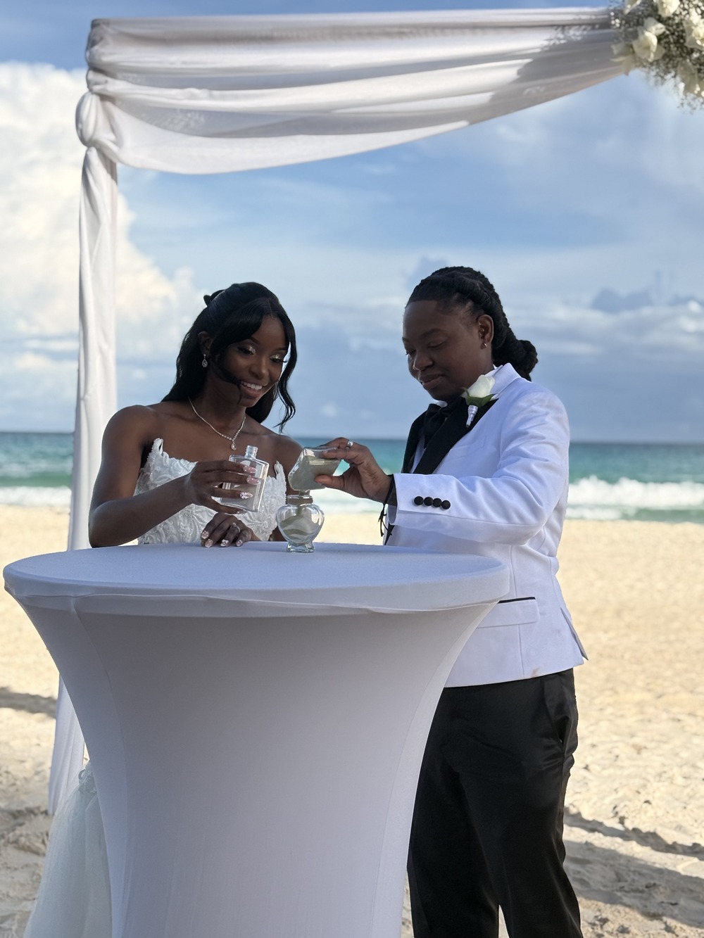 Kathleen and Lucas pour sand at a beach table under an arch, celebrating their destination wedding.