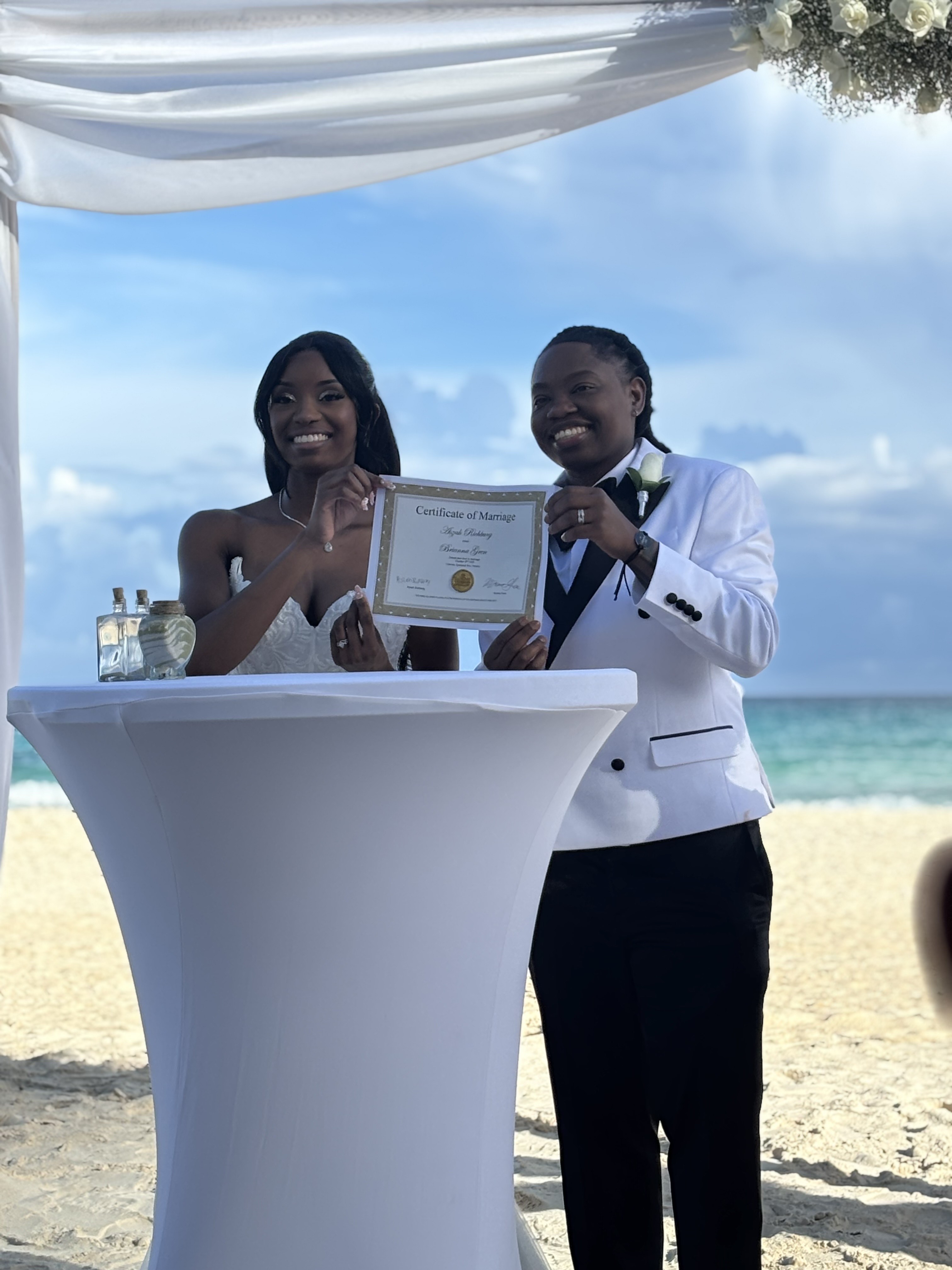 Kathleen and Lucas display their wedding certificate on a beach beneath a white canopy at a destination wedding.