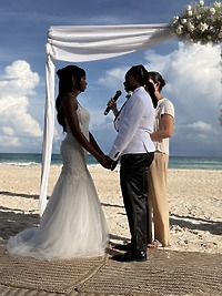 Two brides hold hands beneath a white arch at their destination wedding on the beach.