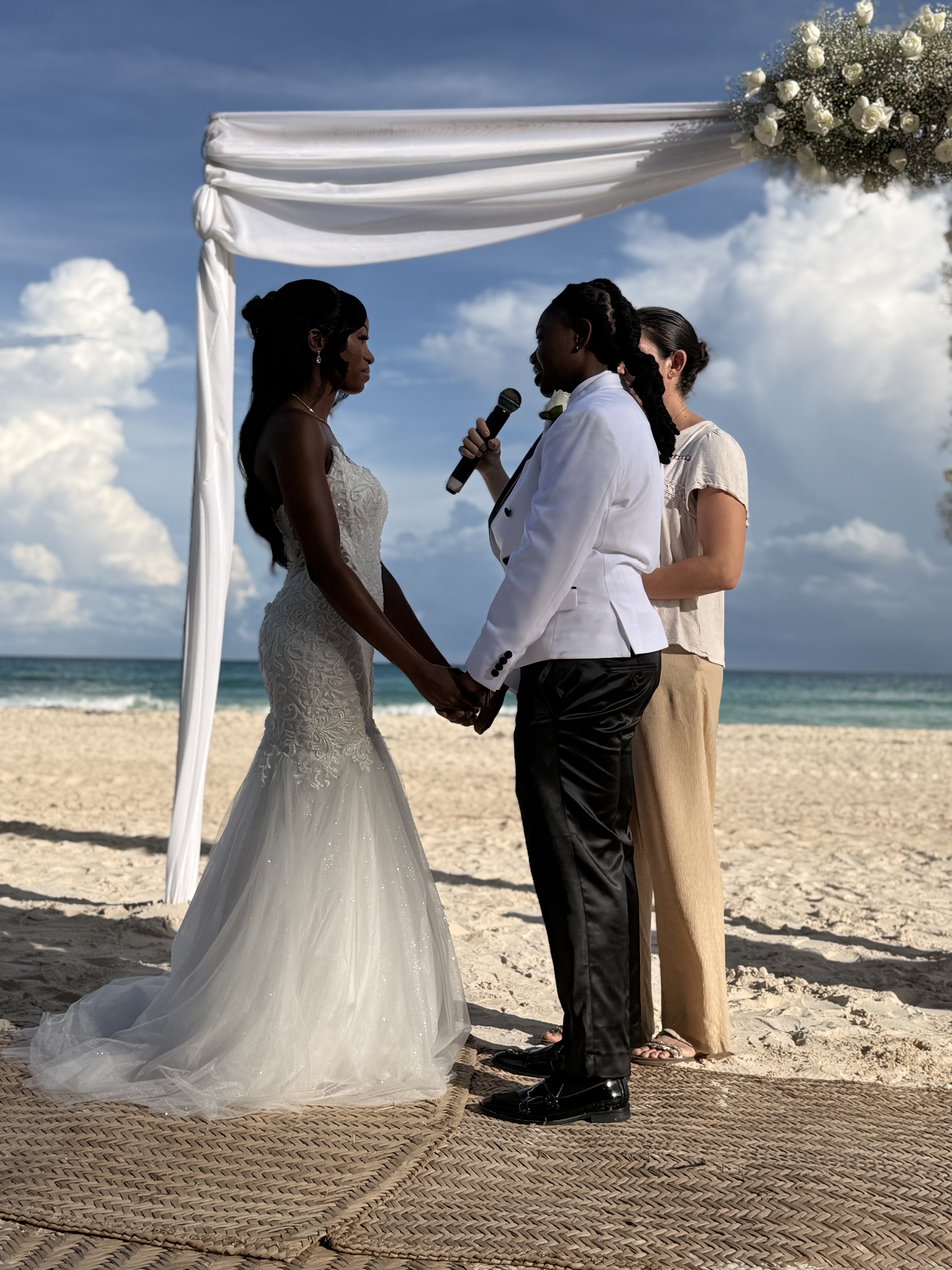Two brides hold hands beneath a white arch at their destination wedding on the beach.