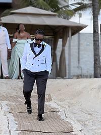 A destination wedding group in formalwear walks a sandy path, led by someone in a tuxedo and sunglasses.