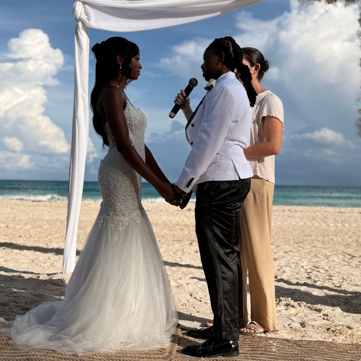 Two brides hold hands beneath a white canopy at their destination wedding. Product: Kathleen and Lucas.