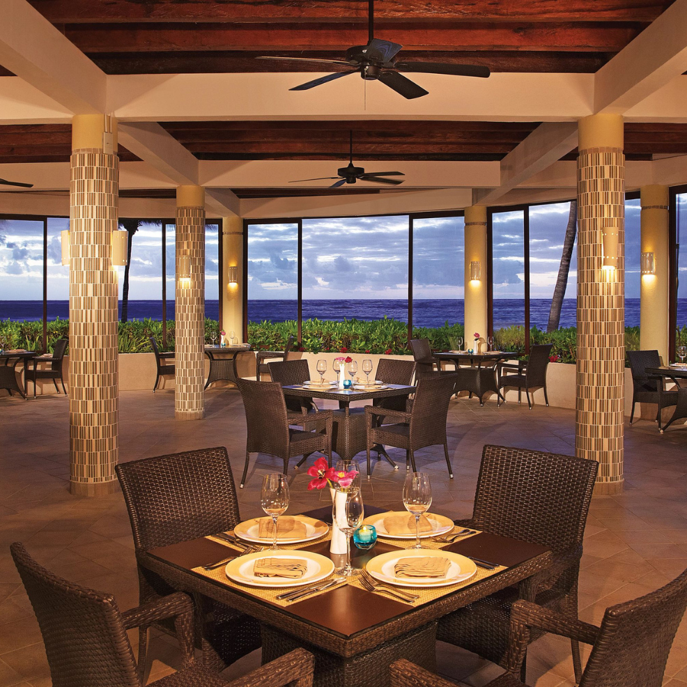 Open-air restaurant set for a destination wedding, with wicker chairs, ocean views, and sunset lighting.