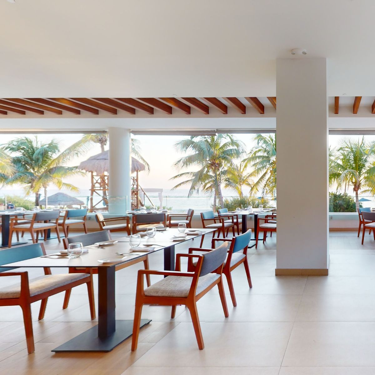 Chic restaurant with wooden chairs and tables, large windows, perfect for a destination wedding; palms outside.