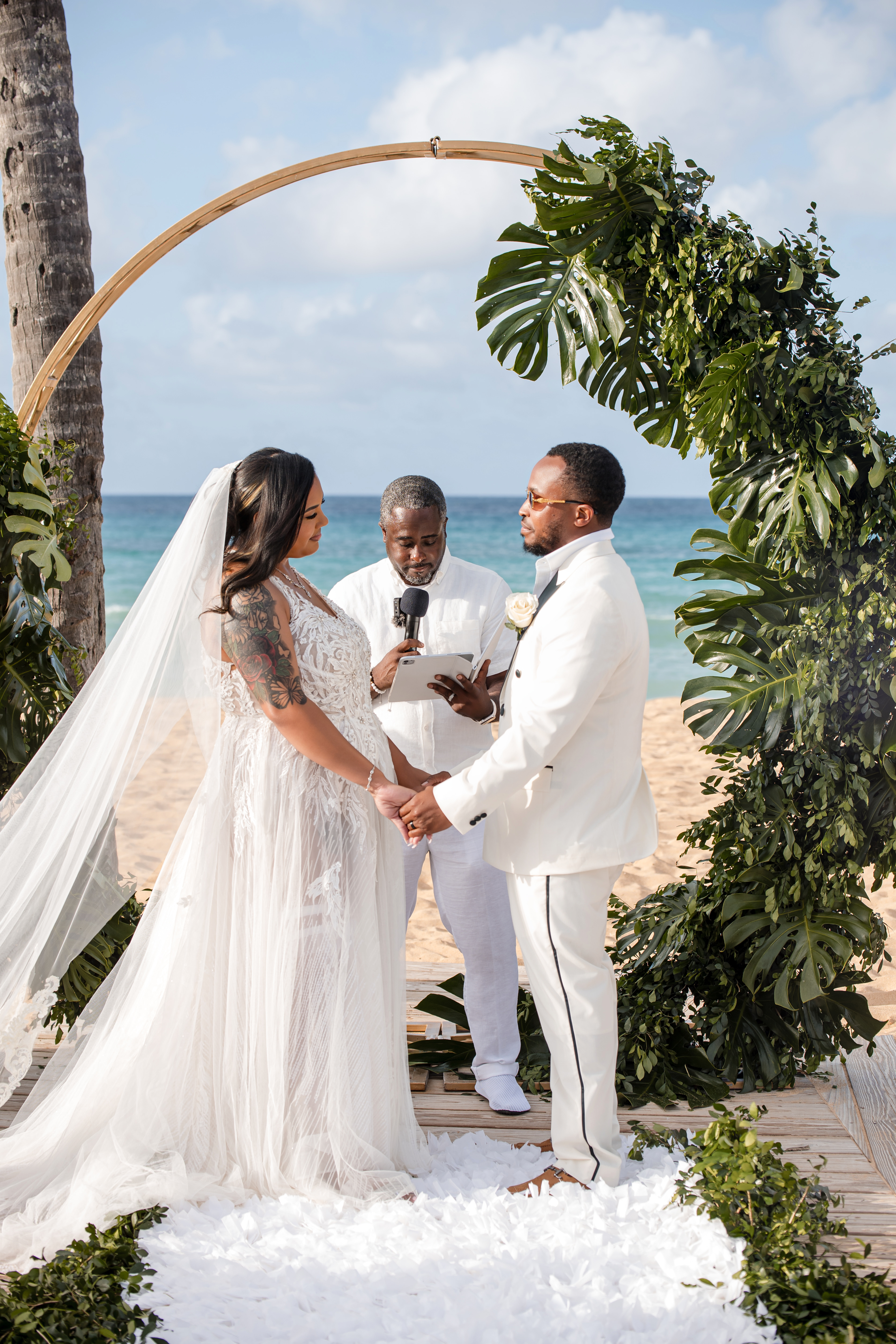 Leah and Ben have a destination wedding, exchanging vows on the beach under a greenery arch with an officiant.