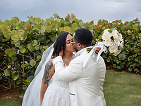 Leah and Ben kissing outdoors in white wedding attire, holding a bouquet near lush green bushes.