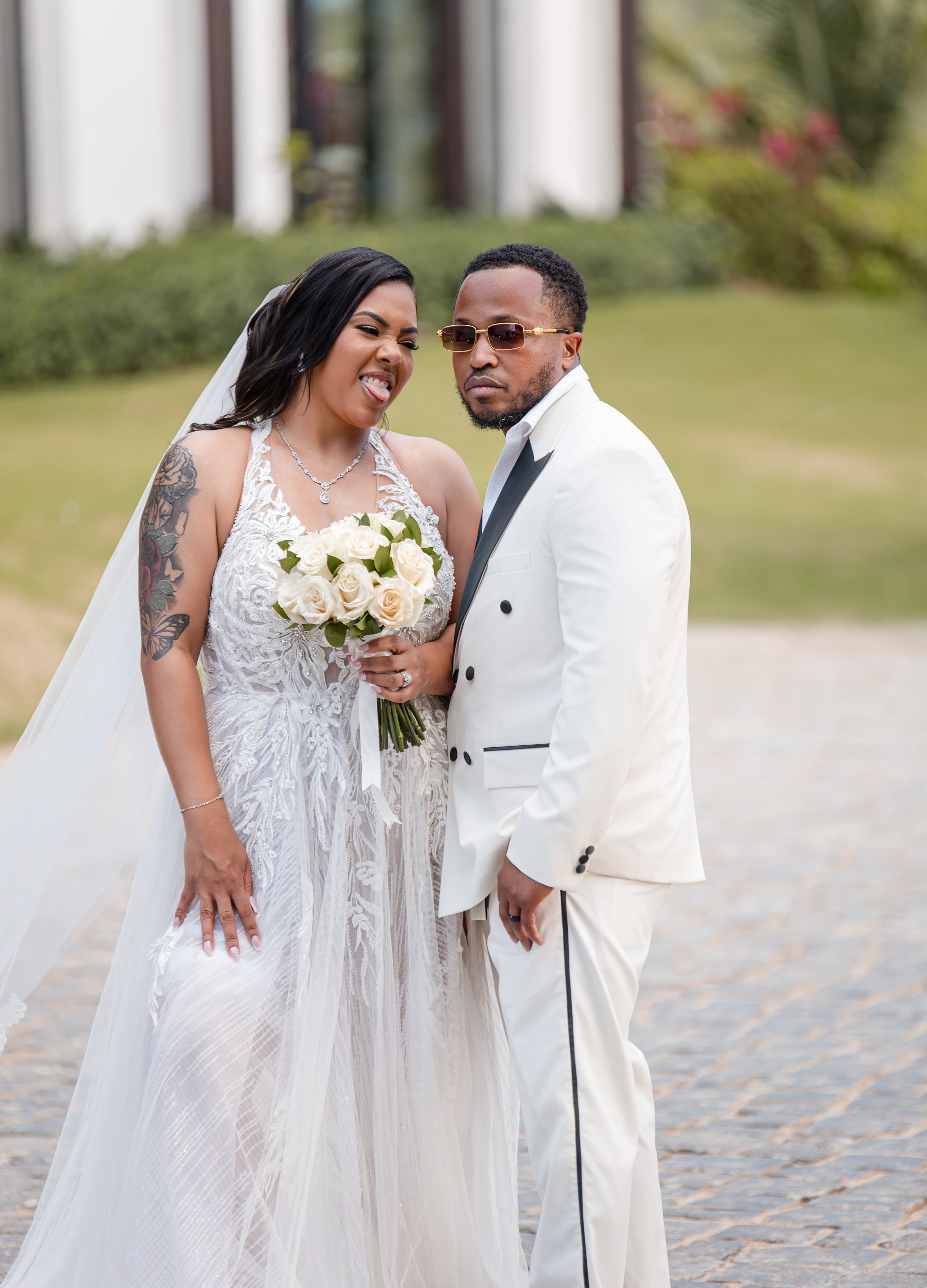 Bride in white dress holding flowers stands with groom in white suit at outdoor destination wedding.