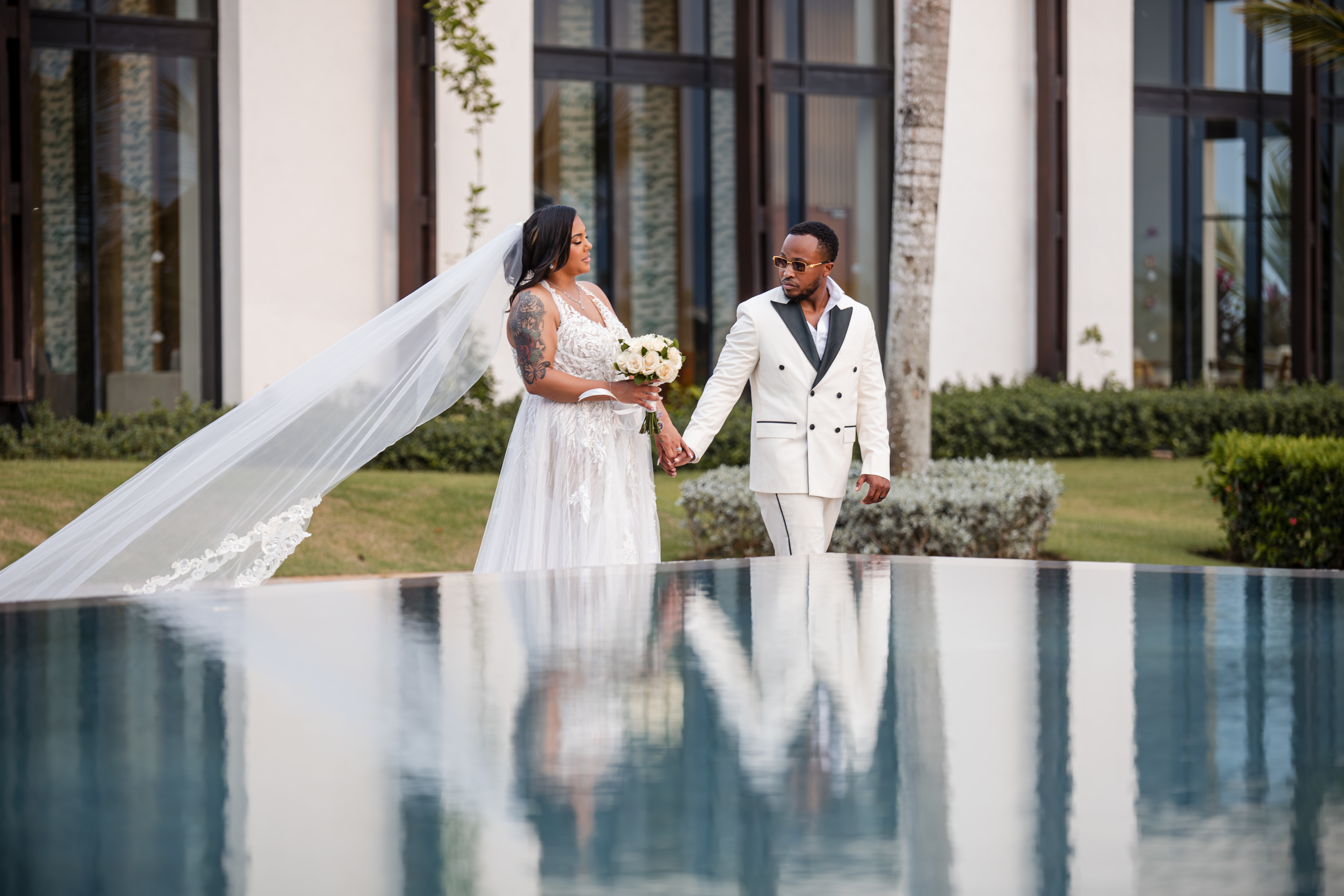 Leah and Ben walk hand in hand by a reflective pool in elegant attire at their destination wedding.