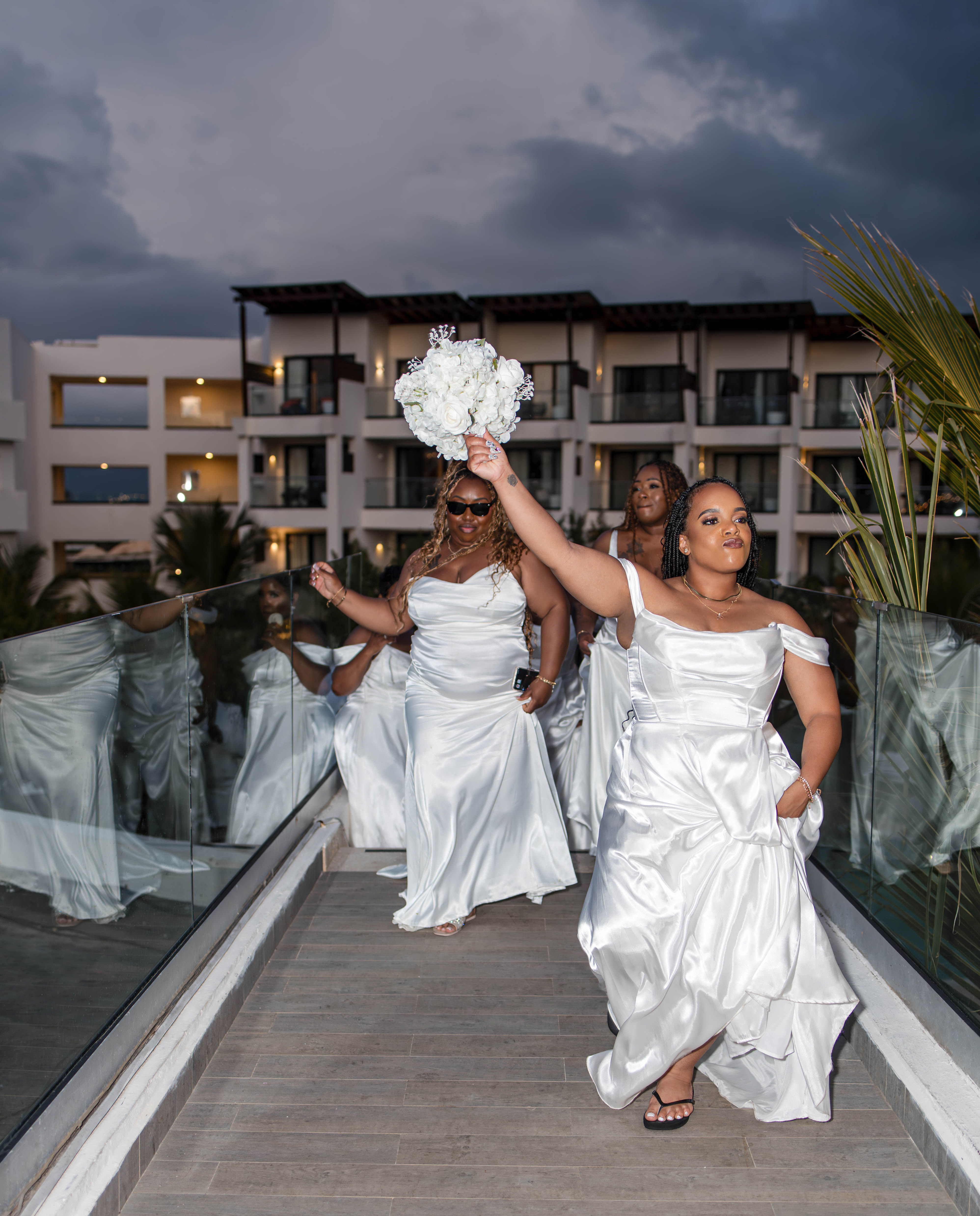 At a destination wedding, four women in white dresses cross a bridge; the leader holds a bouquet.