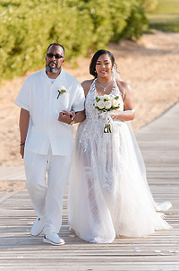 A wedding couple in white walks arm-in-arm on a wooden outdoor path at a destination wedding.