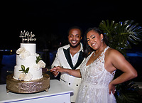 Leah and Ben smile in formal wear as they cut a white wedding cake at a nighttime destination wedding.